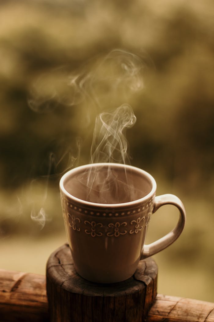A steaming coffee mug sits on a wooden surface, emitting gentle steam in an atmospheric outdoor setting.